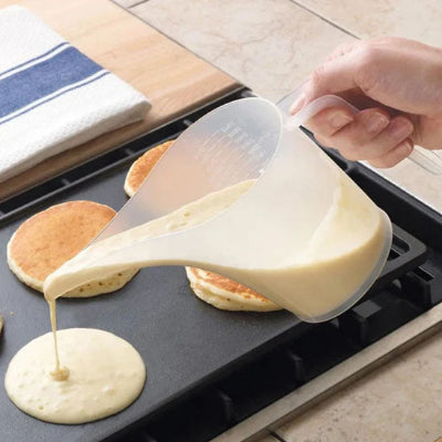 Verre doseur transparent au long bec verseur versant de la pâte à pancakes sur une plaque de cuisson