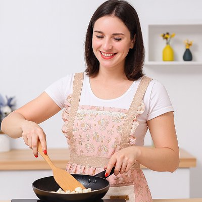 Femme souriante portant tablier de cuisine rose motif floral cuisinant dans poêle avec spatule en bois dans cuisine moderne